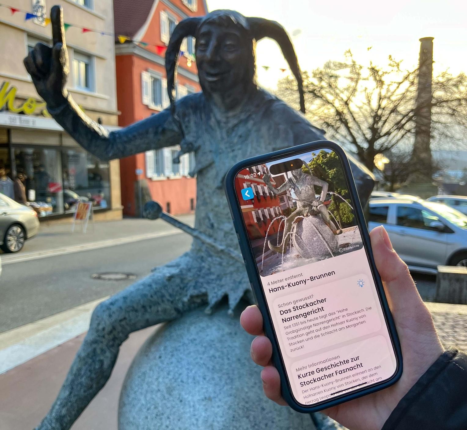 Eine Hand präsentiert ein Handy vor dem Hans-Kuony-Brunnen 